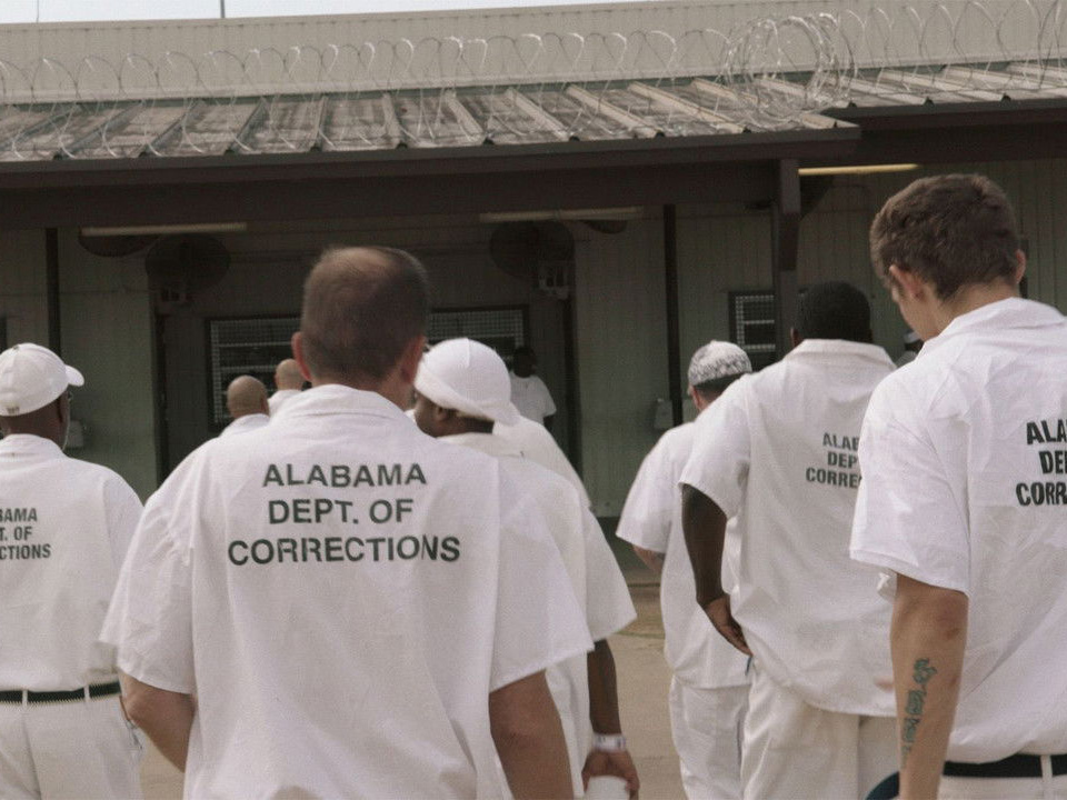 A group of inmates wearing white uniforms with 'Alabama Dept. of Corrections' written on the back, walking across a prison yard towards a building